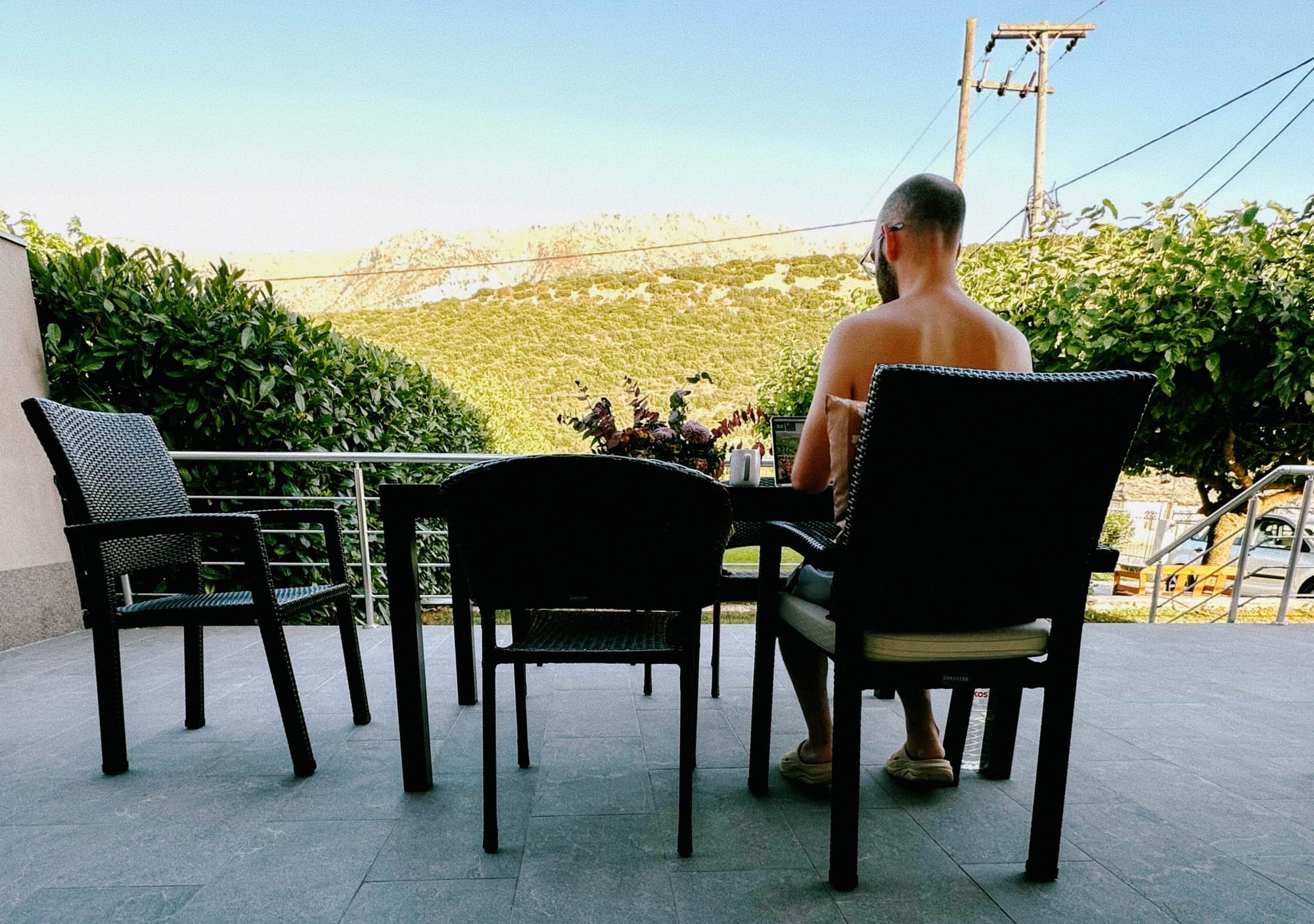 Ein Gründer mit Glatze genießt seine Klarheit und arbeitet an einem Tisch auf der Terrasse mit weitem Blick über die Berge.